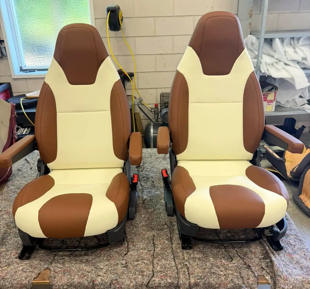 Twee camperstoelen naast elkaar in de werkplaats, opnieuw bekleed in crème leer met bruine zijpanelen en hoofdsteun.